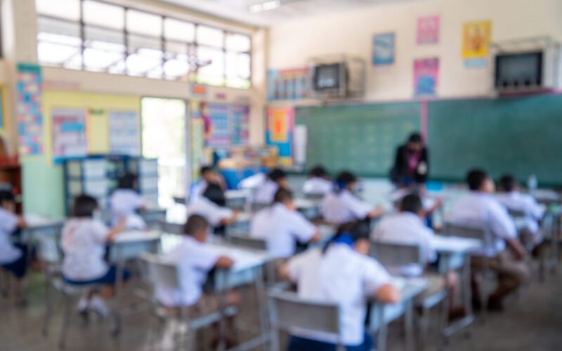 Education evaluation, Blurred image of Writing test in exam with behind child asian students group concentrate in primary school, final examination desk at classroom with Thai student uniform.