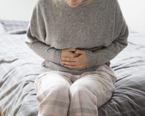 Sick woman in grey homewear sitting on bed, keeping hands on stomach, suffering from intense pain. Illness, stomach ache concept