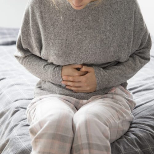 Sick woman in grey homewear sitting on bed, keeping hands on stomach, suffering from intense pain. Illness, stomach ache concept