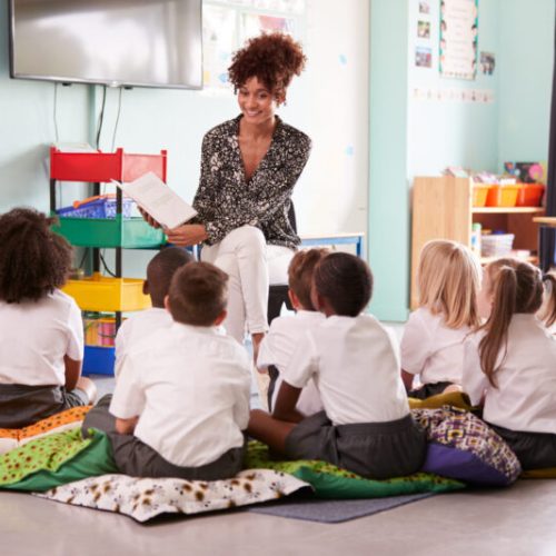 Female Teacher Reading Story To Group Of Elementary Pupils Wearing Uniform In School Classroom