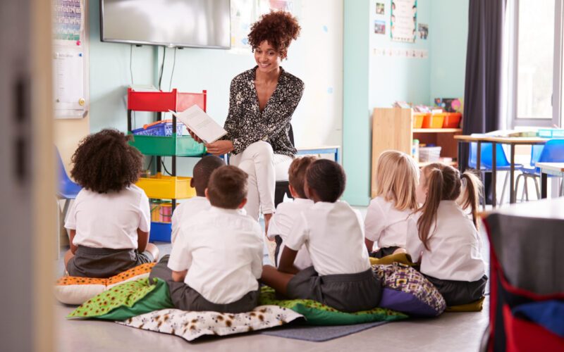Female Teacher Reading Story To Group Of Elementary Pupils Wearing Uniform In School Classroom