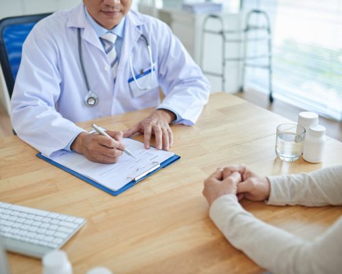 Close-up shot of male doctor filling in medical record, his patient sitting opposite him