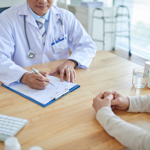 Close-up shot of male doctor filling in medical record, his patient sitting opposite him