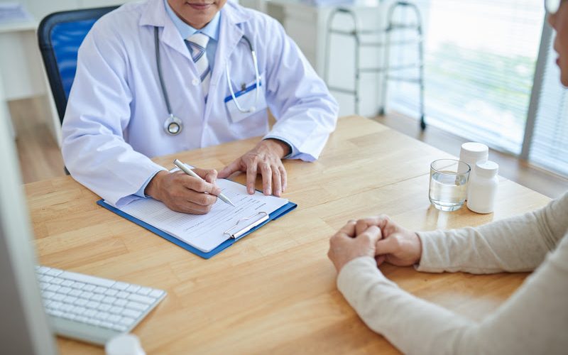 Close-up shot of male doctor filling in medical record, his patient sitting opposite him