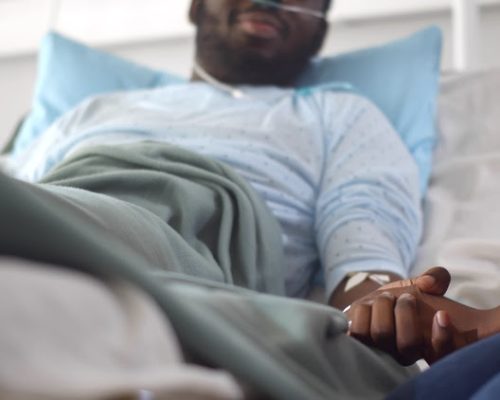 Cropped shot of african wife visiting husband in hospital holding hands and supporting. Sick young black male patient lying in hospital bed and talking to woman visitor in ward