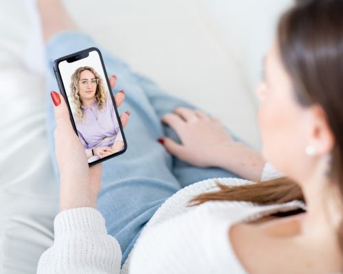 Woman patient listens to careful lady psychologist advice at videocall via mobile phone sitting on sofa in living room close view