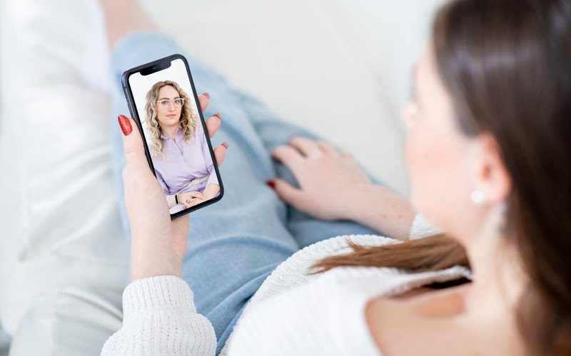 Woman patient listens to careful lady psychologist advice at videocall via mobile phone sitting on sofa in living room close view
