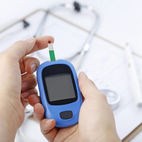 Hand holding a blood glucose meter measuring blood sugar, the background is a stethoscope and chart file