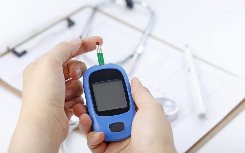 Hand holding a blood glucose meter measuring blood sugar, the background is a stethoscope and chart file